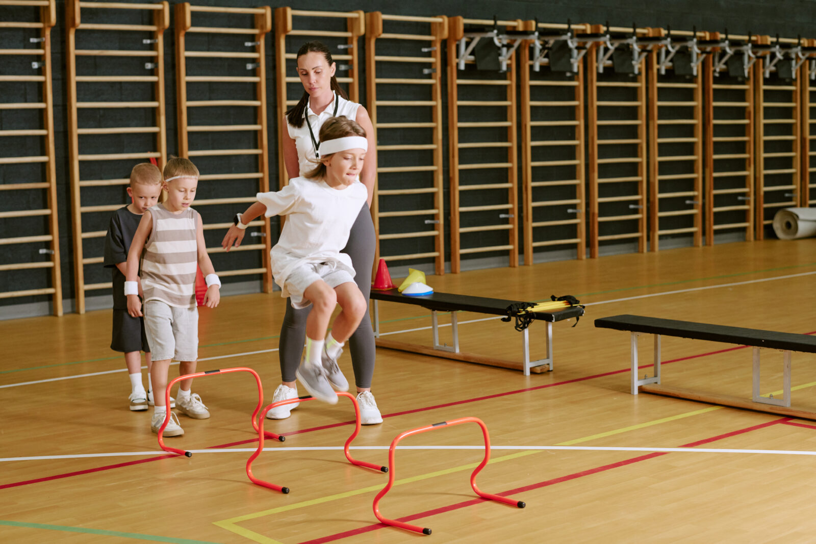 Female Trainer Helping Children Jumping over Hurdles in Gym